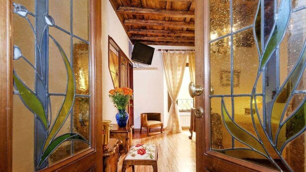 A narrow hallway with wooden doors and a wooden ceiling. There is a small table with a vase of flowers, a chair, and a wooden bench. - Apartment (Roma)