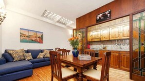 A living room with a blue sofa, wooden dining table, and chairs. There is a kitchen with wooden cabinets and a map on the wall. - Apartment (Roma)