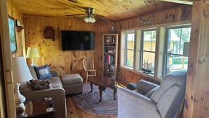 L’intérieur chaleureux d’un chalet avec des murs en bois, un ventilateur de plafond, un téléviseur à écran plat, une bibliothèque et une grande fenêtre.