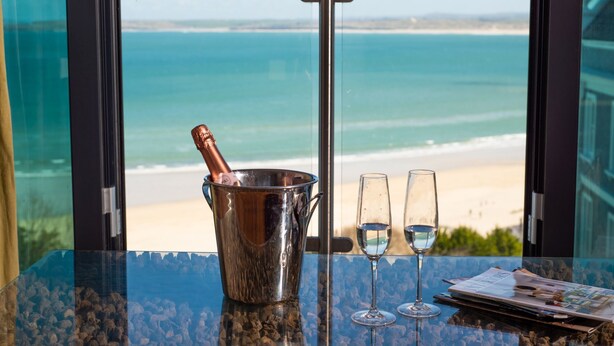 A table with a champagne bottle in a metal bucket, two glasses of champagne, and a magazine on a balcony overlooking a beach.