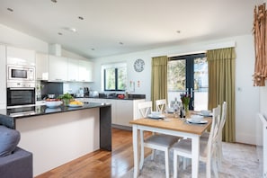 A modern kitchen with built-in appliances, a dining area with a wooden table and white chairs, and a large window with curtains.