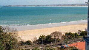 A beach with a tennis court in the foreground, a sandy beach, and a view of the sea and distant hills.