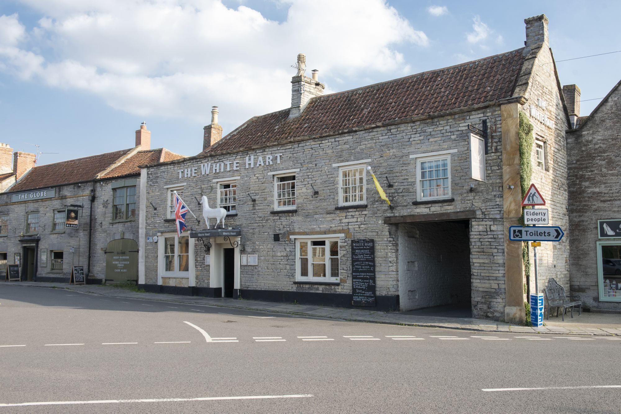 Un bâtiment traditionnel en pierre, sur lequel on peut voir une enseigne indiquant le pub «The White Hart », avec un drapeau, une statue de cheval blanc et un panneau affichant les plats proposés.