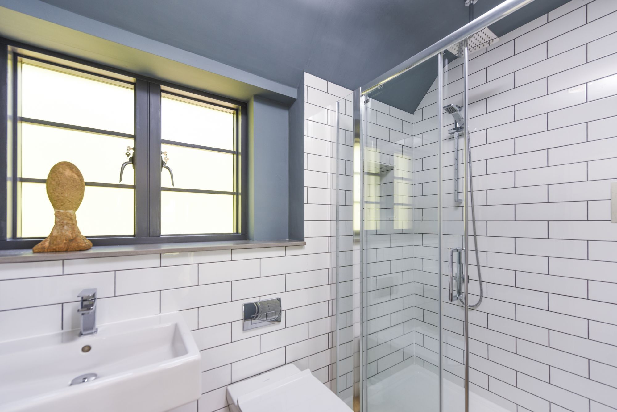 Et moderne badeværelse med en glasbrusekabine, en vask med vandhane og et vindue med en pynteting på gerigten.