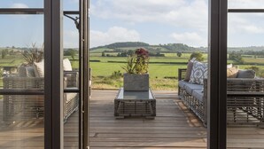 Un patio in legno con mobili da esterno, una fioriera con piante e una vista panoramica su campi e colline verdi.