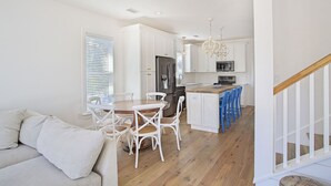 A modern kitchen with white cabinetry, a central island, and a dining area with white chairs. - Periwinkle - Sunrise Beach on 30A, Heated Private Pool, Free Beach Service! (Santa Rosa Beach)