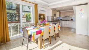 A dining area with a table set for four, a kitchen in the background, and a balcony with a view of greenery. - 4 Ensuite Bedrooms Private Heated Outdoor Pool (Mugla)