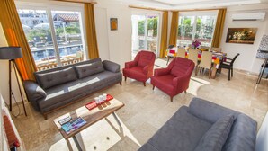 A spacious living room with a grey sofa, red armchairs, and a coffee table with magazines. A balcony with a dining area and large windows is visible. - 4 Ensuite Bedrooms Private Heated Outdoor Pool (Mugla)
