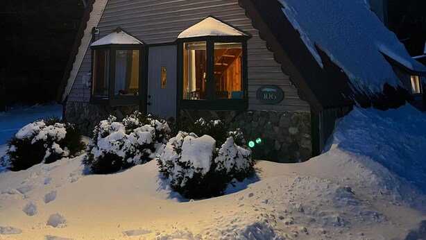 Een huis met een steil dak, een schoorsteen en een verlicht raam. Er ligt sneeuw op het dak en de omliggende struiken.
