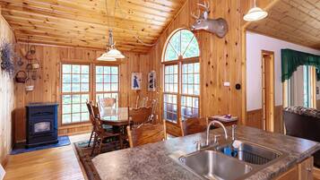 L’intérieur d’un chalet rustique est agrémenté d’une table à manger en bois, de chaises, d’une cheminée en pierre et d’une cuisine dotée d’un plan de travail et d’un évier en granit.