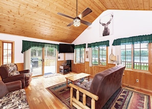L’intérieur chaleureux d’un chalet avec un plafond en bois, un ventilateur de plafond, une grande fenêtre donnant sur un lac et un téléviseur fixé au mur.