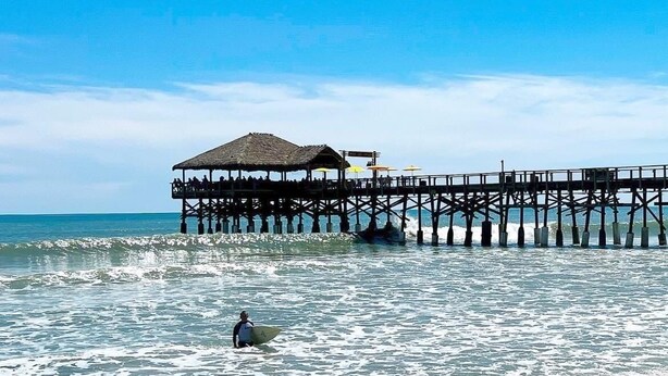 A beach with a pier extending into the ocean, featuring a thatched-roof structure with seating.