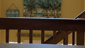 A room with a large window overlooking a tropical scene with palm trees and a view of the ocean. There is a framed picture of palm trees on the wall below the window.