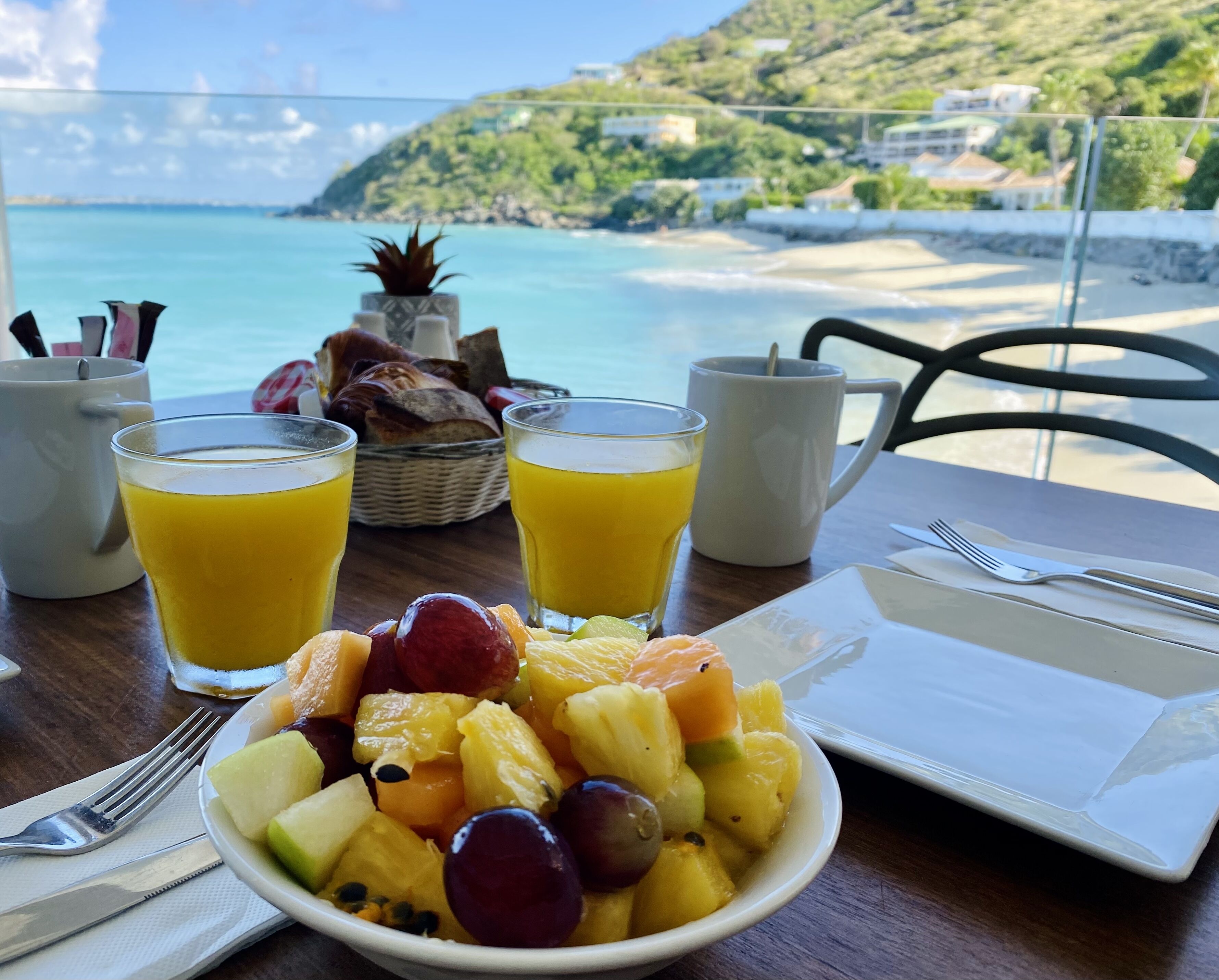 Una mesa con frutero, dos vasos de jugo de naranja, un plato y una taza, con vista a una playa y colinas.