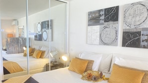 Une chambre d’hôtel moderne avec un grand lit, du linge de lit blanc et des coussins décoratifs. On y trouve également des œuvres d’art murales et un miroir qui reflète la pièce.