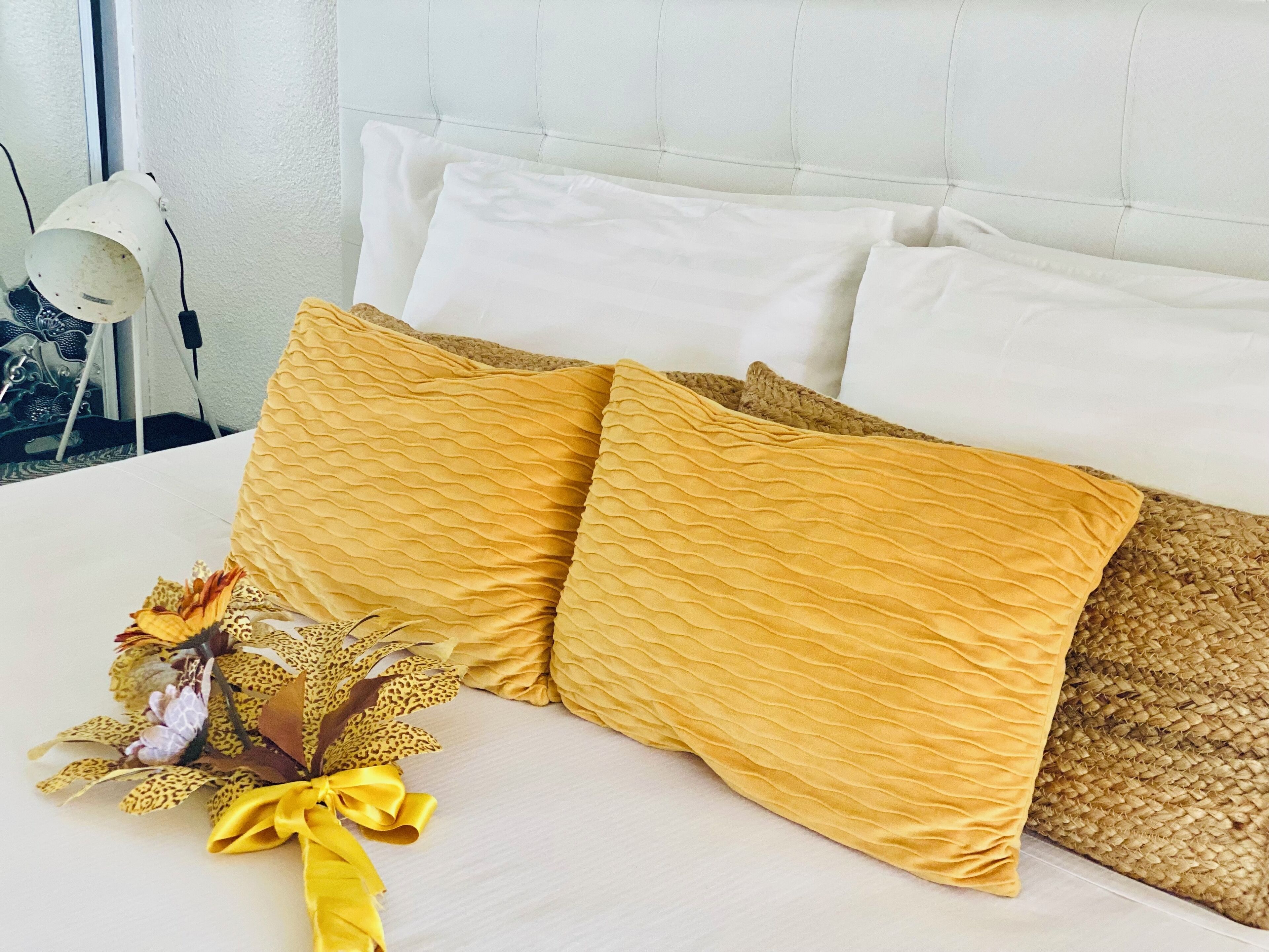 Una cama bien hecha con almohadas blancas y amarillas, un arreglo floral decorativo y una lámpara en la mesita de noche.