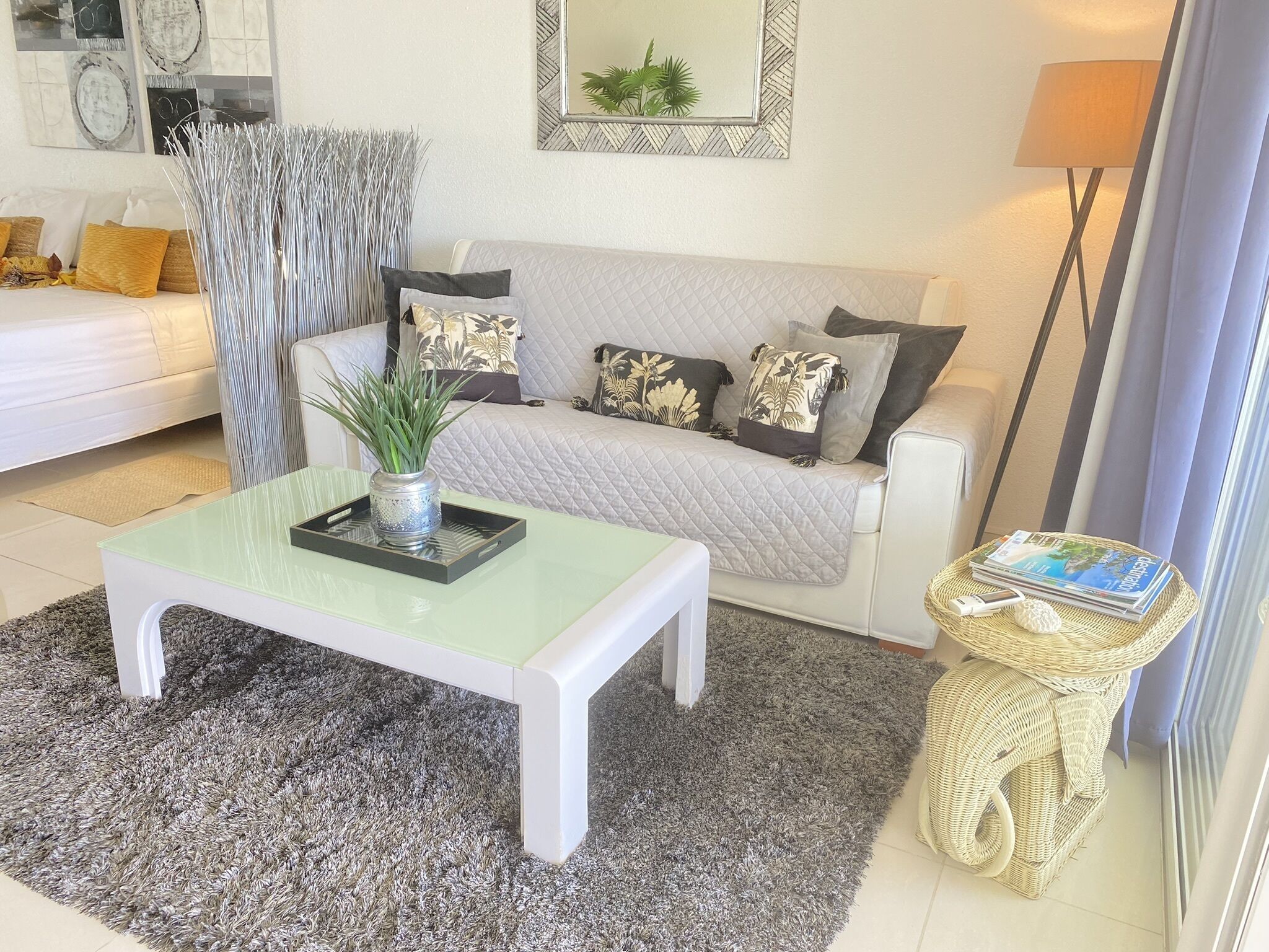 A living room with a white sofa, a glass coffee table, a grey rug, and a wicker side table with magazines.