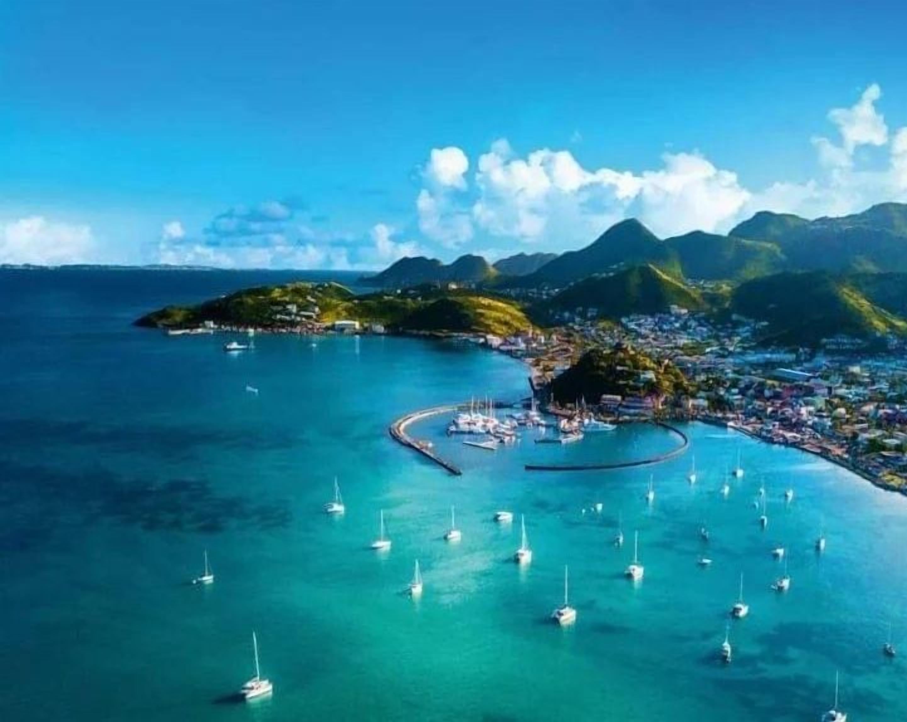 Une ville côtière dotée d’un port de plaisance où sont amarrés de nombreux voiliers, entourée de montagnes et bordée d’eaux d’un bleu limpide.