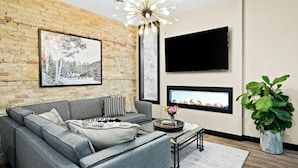 Una sala moderna con un sofá seccional gris, un televisor de pantalla plana en la pared, una chimenea y una planta grande en maceta.
