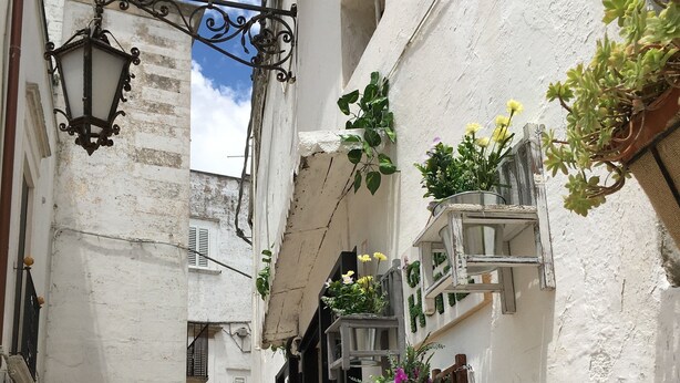 Une ruelle étroite bordée de bâtiments blancs, avec une porte noire et des pots de fleurs suspendus.