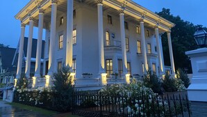 A large, white building with columns and a balcony, surrounded by a black iron fence and white flowers.