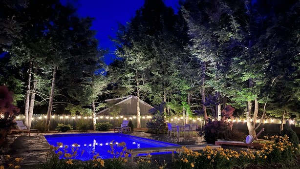A swimming pool surrounded by trees and outdoor furniture, with string lights along the fence.