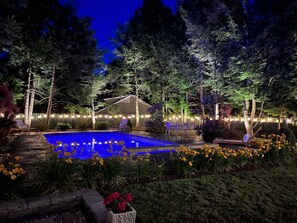Une piscine entourée d’arbres et agrémentée de mobilier d’extérieur, avec des guirlandes lumineuses le long de la clôture.