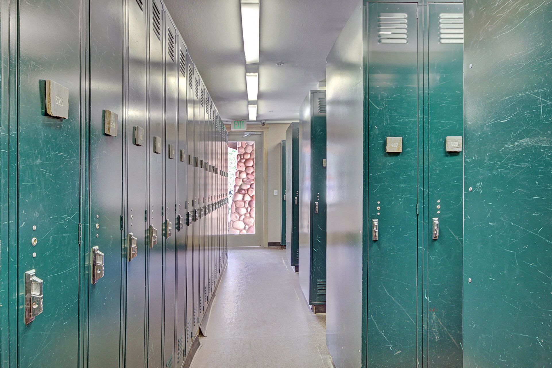 A hallway with rows of lockers, a stone wall, and an exit sign.
