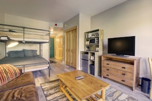 Un espacio habitable compacto con literas, una mesa de madera, un televisor, un microondas y una pequeña zona de cocina.