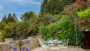 Un patio al aire libre con sillas y una mesa de metal blanco, rodeado de follaje y un muro de piedra.