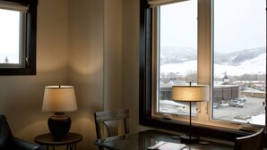 A room with a dining table, chairs, a lamp, and a window with a view of a snowy landscape and buildings. - Ice Skating Suite 202 at Granby Dairy Suites (Granby)