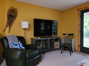 A living room with a black leather sofa, a television on a grey stand, a floor lamp, and a window with a view of greenery. - Snowbrook: Ski in/out access with pool, hot-tub, and sauna for après ski! (Carrabassett Valley)