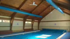 Una piscina interior con vigas de madera, ventiladores de techo y carteles que indican que no se permite bucear.