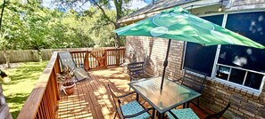 Una terraza de madera con una mesa y sillas de vidrio, una sombrilla verde y un muro de ladrillo.