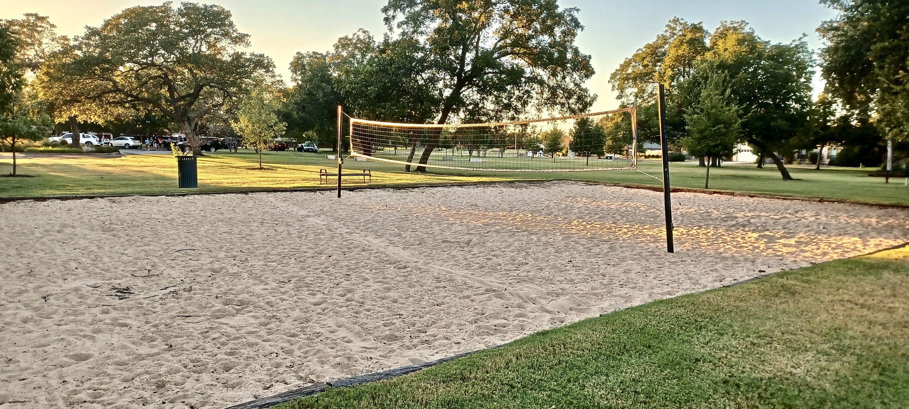 Una cancha de voleibol de arena con una red, rodeada de césped y árboles.