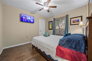 Ein Schlafzimmer mit einem Bett, einem Deckenventilator, einem an der Wand montierten Fernseher, einem Fenster mit Vorhängen und einem gerahmten Bild.