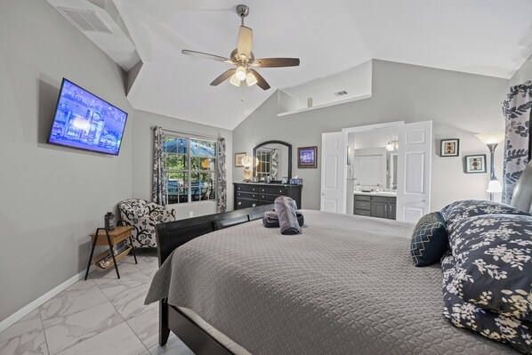 A bedroom with a bed, a desk, a television, a ceiling fan, and a bathroom visible through an open door.