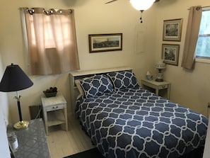 Um quarto com cama, mesa de cabeceira, abajur, janela com cortinas e quadros na parede.