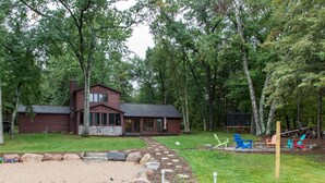 A cabin with a stone foundation, surrounded by a forest. There is a gravel path leading to the cabin, and outdoor seating with chairs. - Private Beach, Beautiful Lake Home, Fishing, Recreation - Everything You Need! (Crosslake)