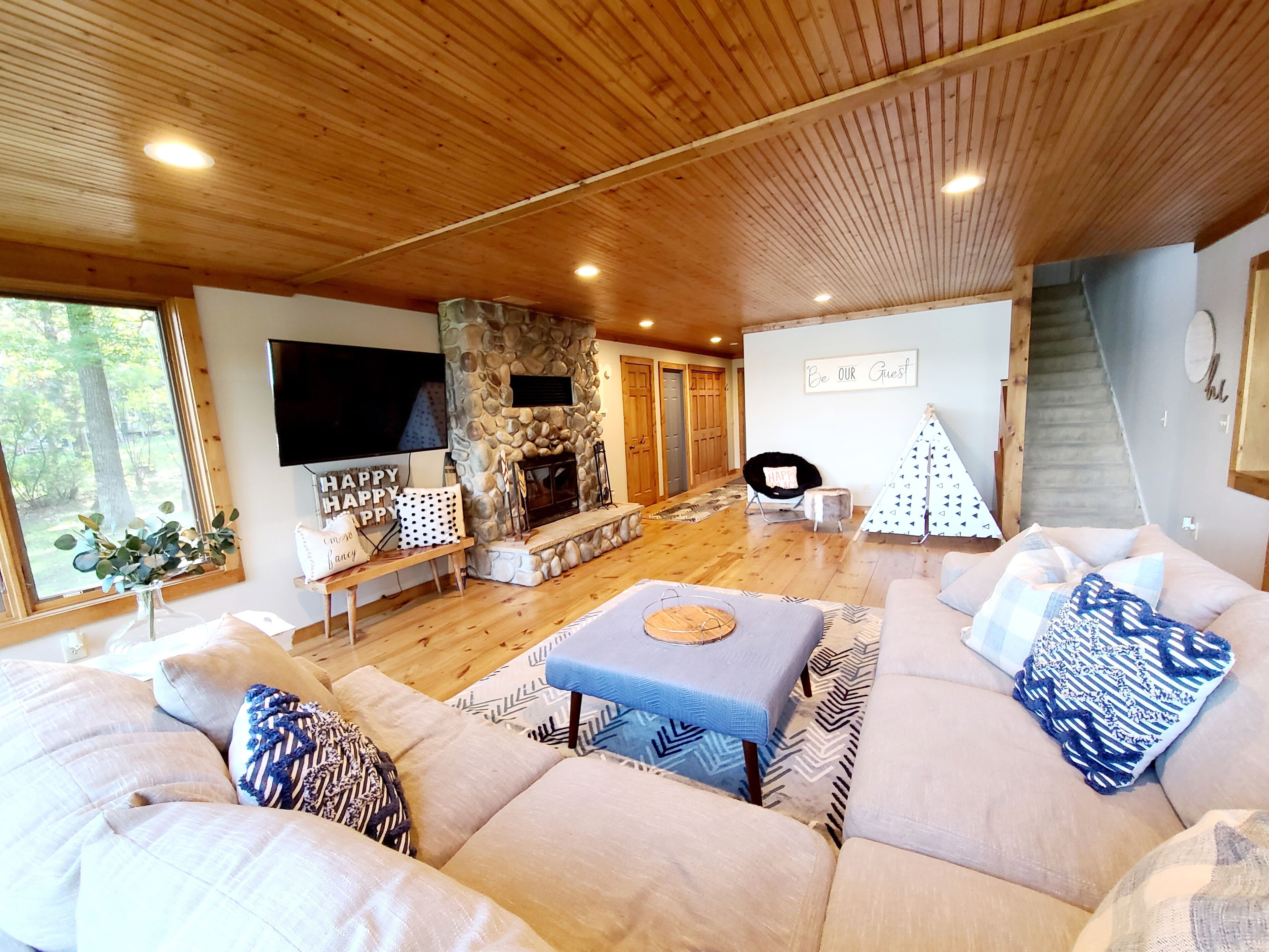 A cozy living room with a stone fireplace, wooden ceiling, and a large window overlooking a forest.