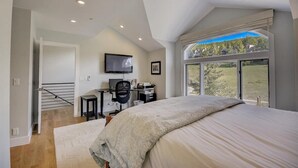 A bedroom with a large bed, a desk with a computer, and a TV. There is a window with a view of trees and a house.