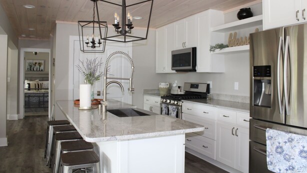 Una cocina moderna con gabinetes blancos, electrodomésticos de acero inoxidable, una isla central con taburetes e iluminación colgante.