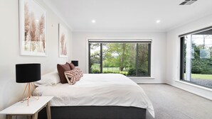 A modern bedroom with a large bed, a bedside table with a lamp, and a view of greenery through large windows.