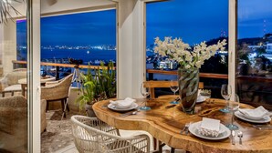 Uma área de refeições com uma mesa de madeira posta para dois, cadeiras brancas e uma grande janela com vista para a cidade à noite.
