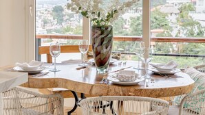 Uma mesa de jantar posta com louça branca e um vaso de flores brancas, cercada por cadeiras de vime, com vista para o horizonte urbano através de grandes janelas.