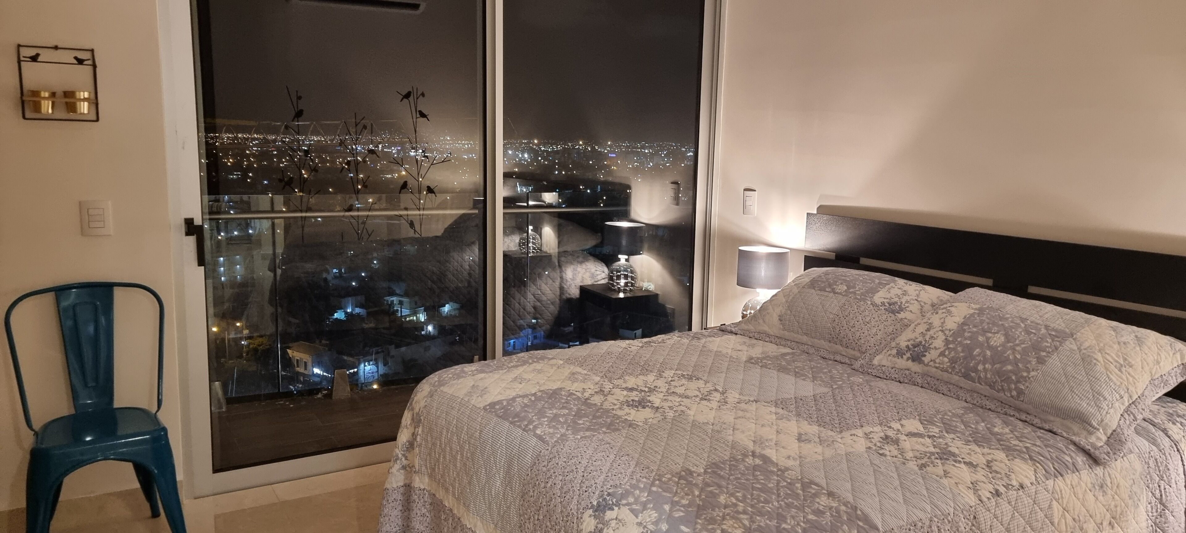 A bedroom with a bed, a chair, a nightstand, and a view of a cityscape at night.