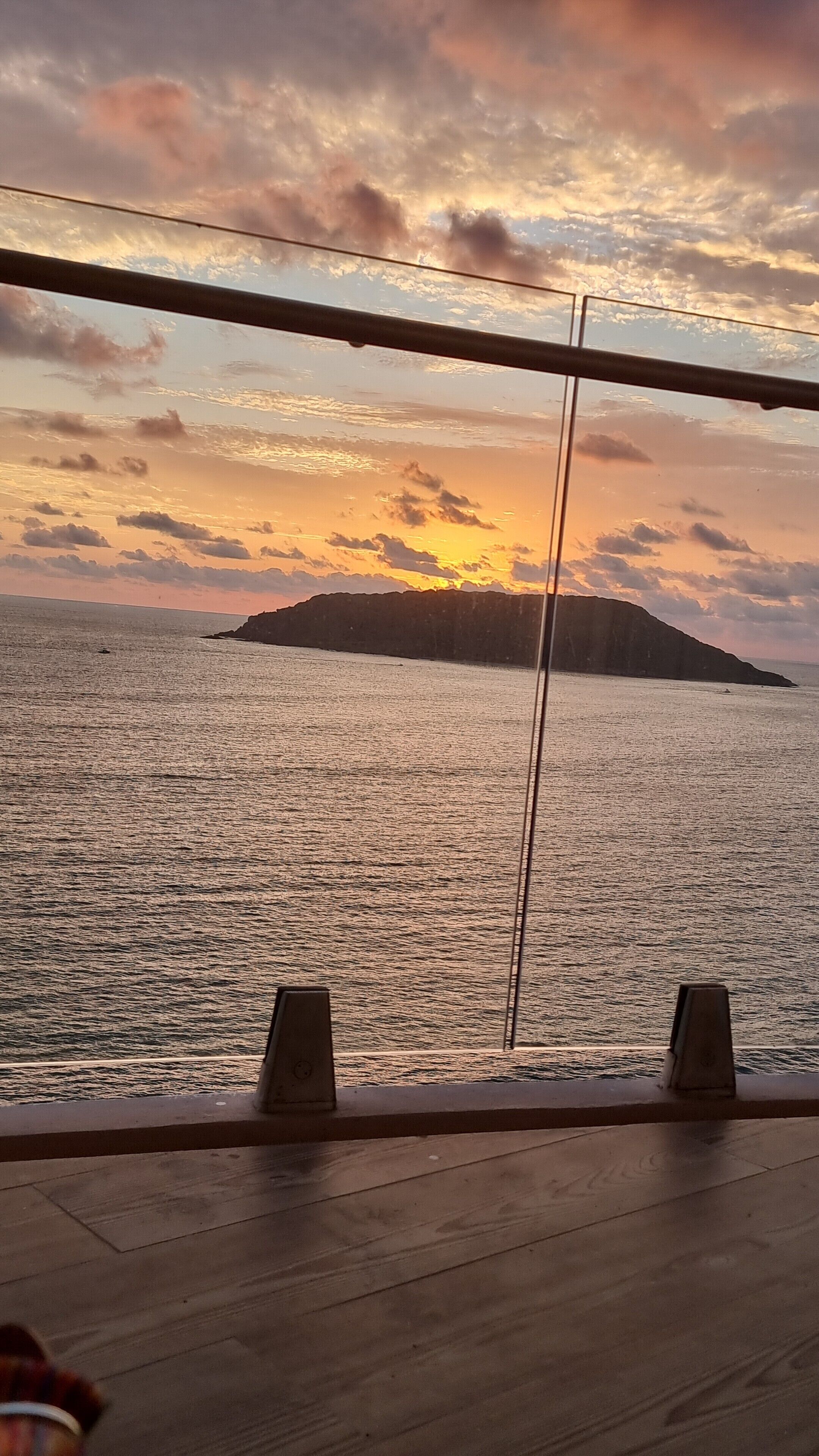 Une vue sur le coucher de soleil depuis un balcon doté d’une balustrade, donnant sur une étendue d’eau et une île lointaine.