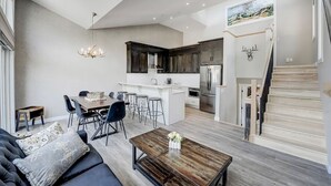 Un salon moderne avec un canapé, une table basse et un coin repas. On aperçoit une cuisine dotée de meubles sombres et d’appareils électroménagers en acier inoxydable. Un escalier mène à l’étage supérieur.