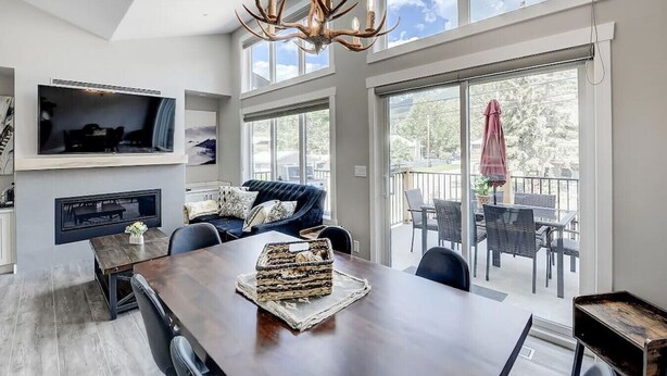 Un salon moderne avec une grande table à manger en bois, un canapé confortable et une cheminée. Il y a un balcon avec des sièges d’extérieur et un téléviseur fixé au mur.