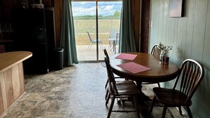 Een eethoek met een houten tafel en stoelen, een keuken met een koelkast, en een uitzicht door een glazen deur op een terras.